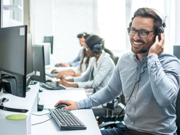 Smiling man with headset at computer desk answering phones, others sitting behind him doing the same