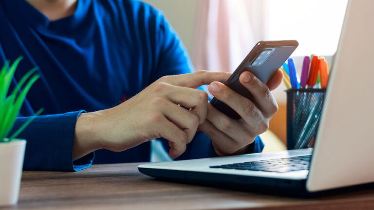 A person is sitting at a desk with a laptop computer and searching on their mobile phone.