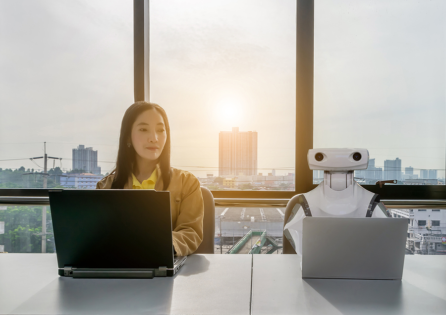 A woman and a robot sit side-by-side at a desk in an office with laptop computers.