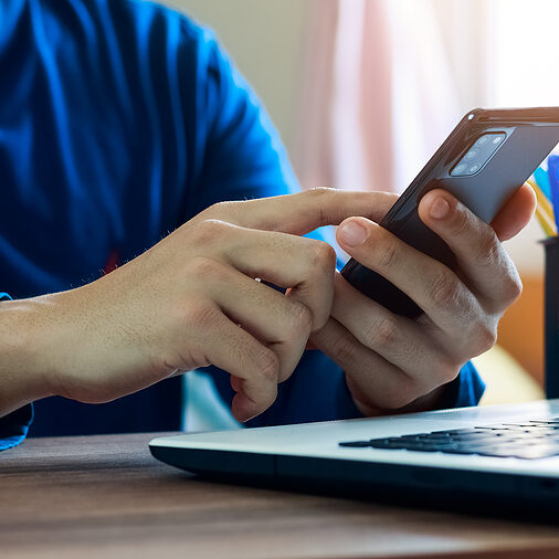 A person is sitting at a desk with a laptop computer and searching on their mobile phone.