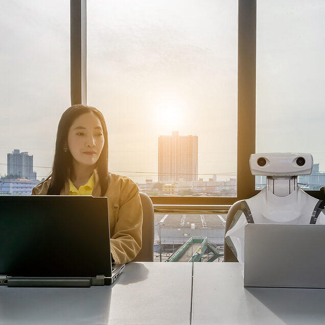 A woman and a robot sit side-by-side at a desk in an office with laptop computers.